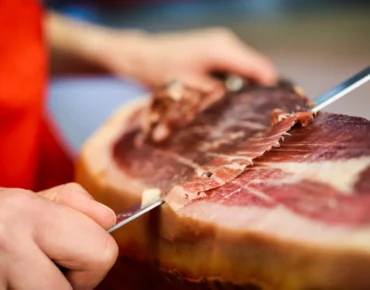 Mano cortando jamón curado con un cuchillo afilado, mostrando la textura y el color de la carne. Fondo desenfocado que resalta la atención en el proceso de corte.