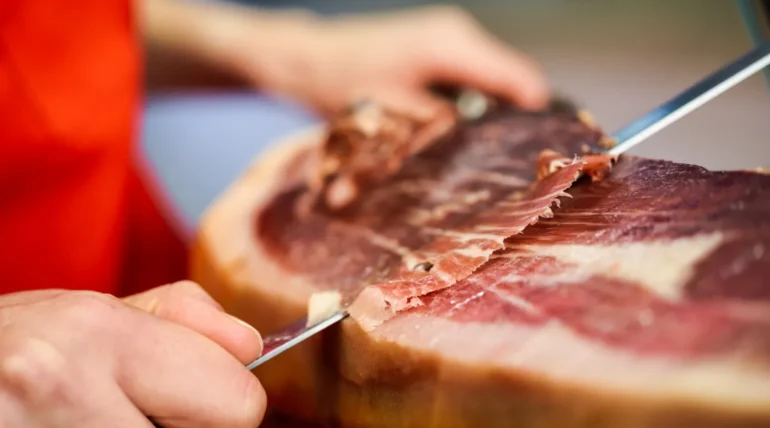 Mano cortando jamón curado con un cuchillo afilado, mostrando la textura y el color de la carne. Fondo desenfocado que resalta la atención en el proceso de corte.