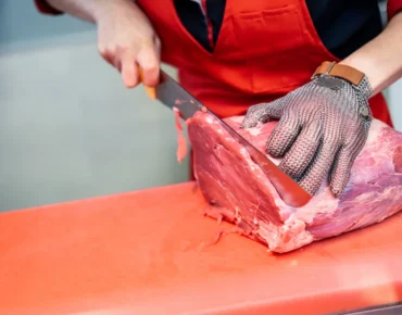 Carnicero cortando carne roja con un cuchillo en una tabla de trabajo, usando guantes de protección y un delantal rojo.