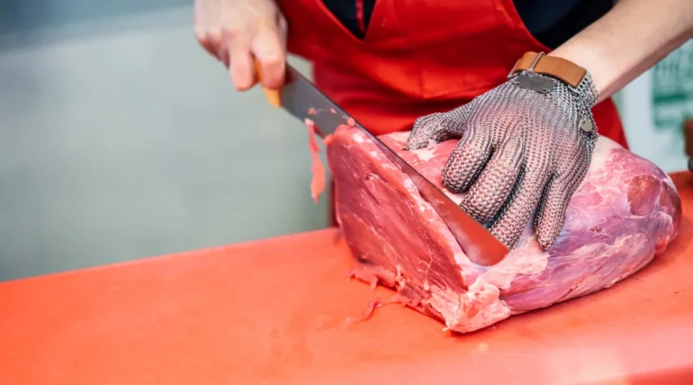 Carnicero cortando carne roja con un cuchillo en una tabla de trabajo, usando guantes de protección y un delantal rojo.