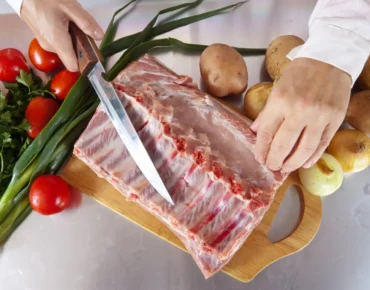Mano cortando costillas de cerdo con un cuchillo en una tabla de madera, rodeada de ingredientes frescos como tomates, cebollas, papas y cilantro. Ideal para recetas de cocina.