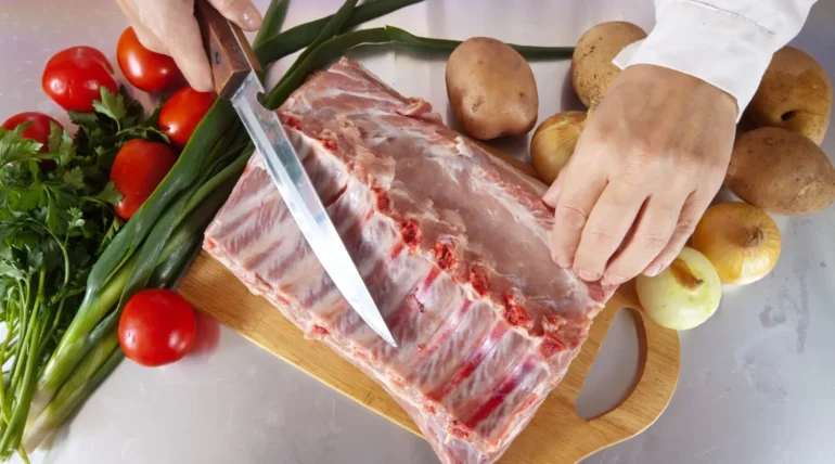 Mano cortando costillas de cerdo con un cuchillo en una tabla de madera, rodeada de ingredientes frescos como tomates, cebollas, papas y cilantro. Ideal para recetas de cocina.